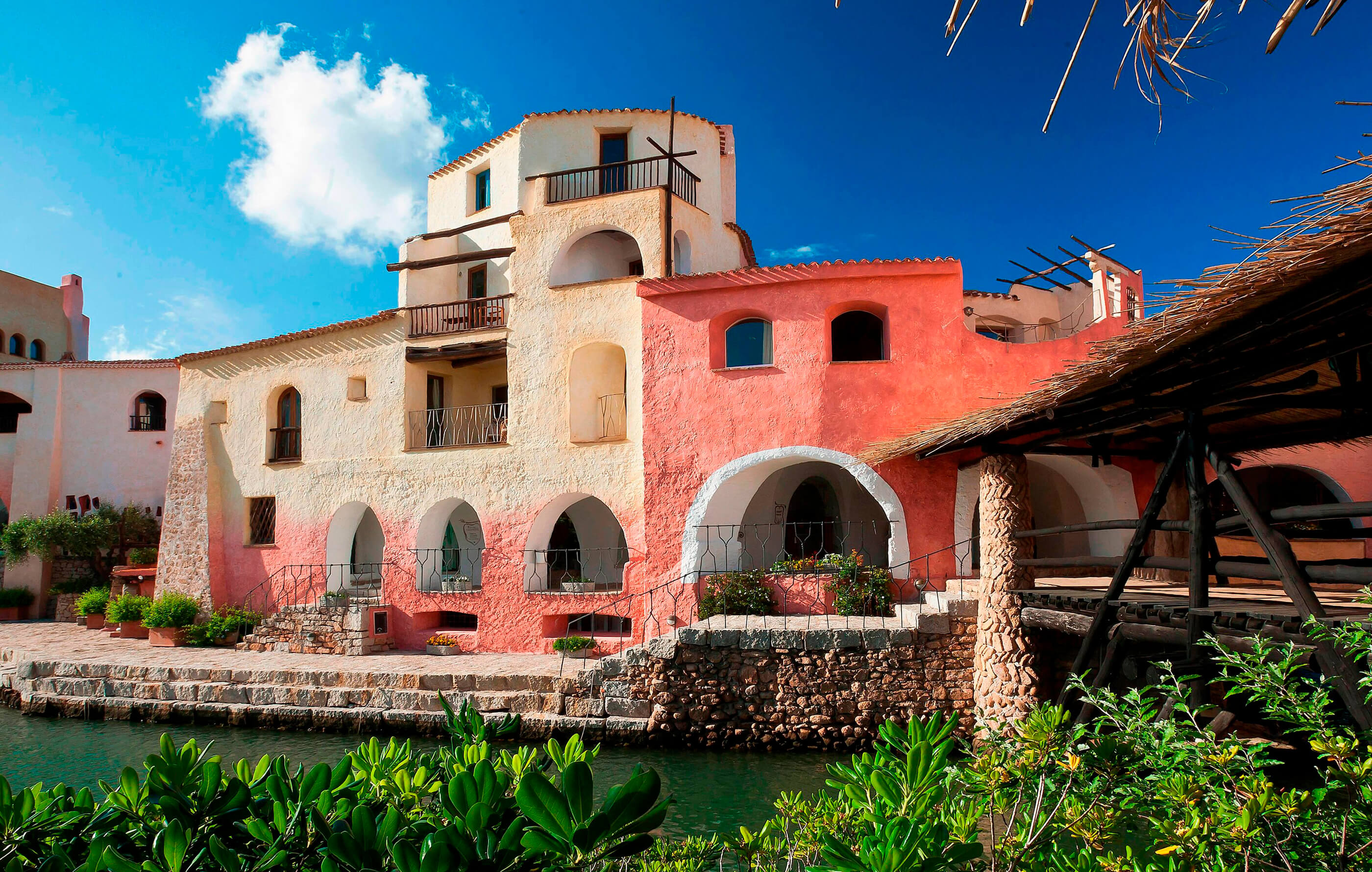 Full screen image of a photo of the Cala Di Volpe Hotel, in Sardinia, with a thatched roof walkway and rough rendered buildings covered in pastel pink and yellow textures