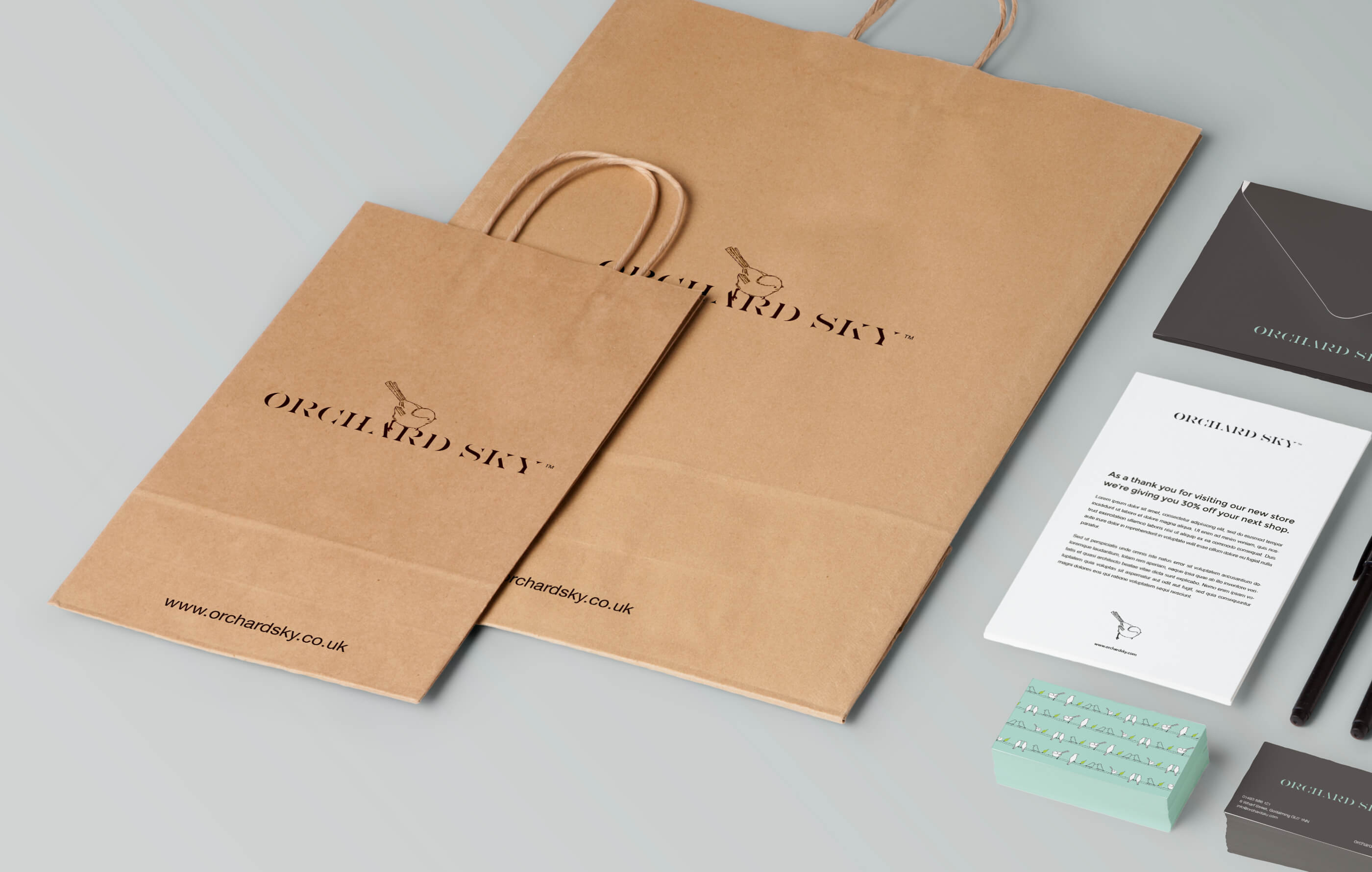 Photorealistic mock up of Orchard Sky stationery and branded collateral, including brown paper bag, business card, pen and an envelope