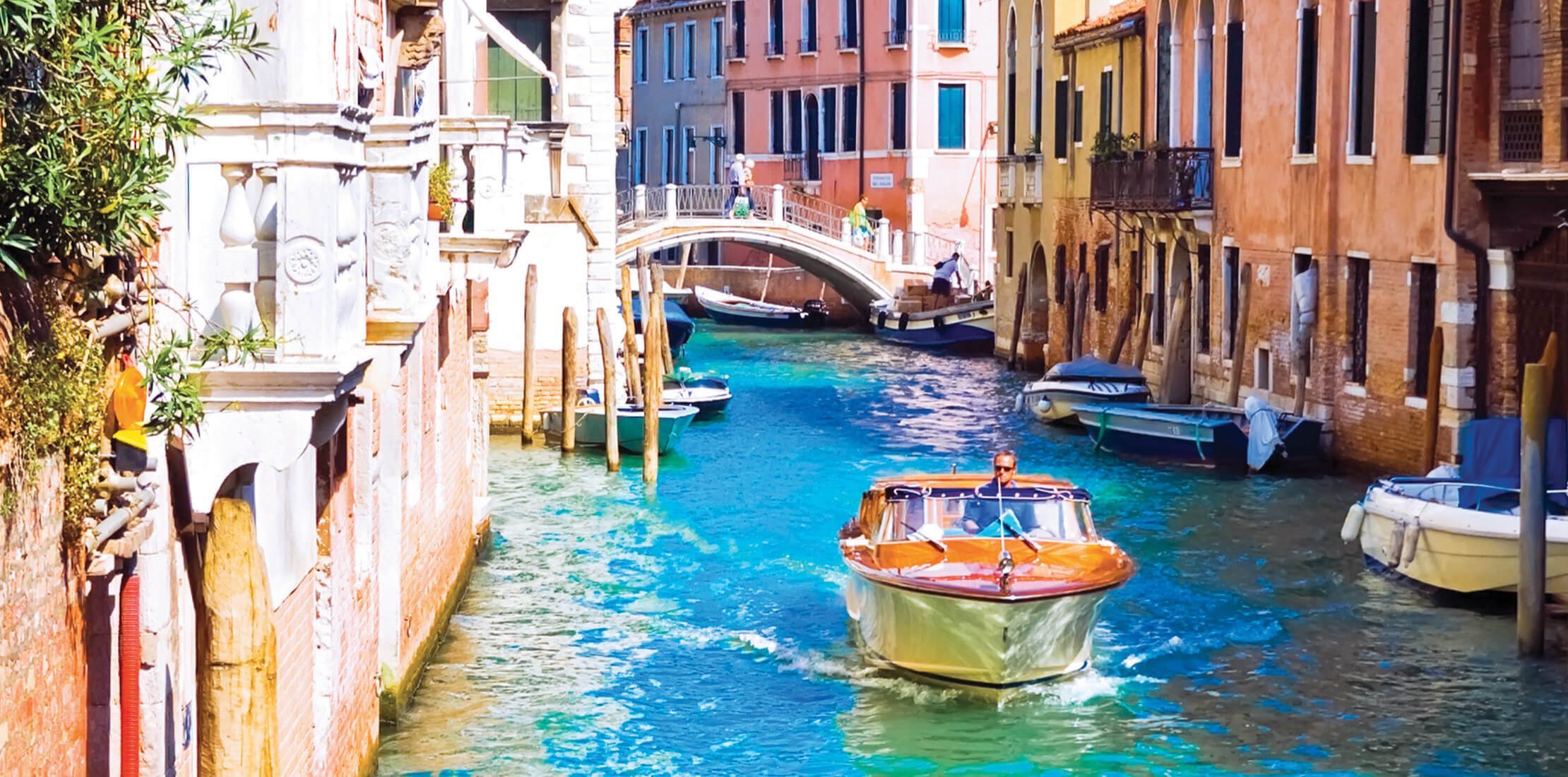 Sunny shot of a canal in Venice, with boats moared along its sides and a small stone bridge in the background
