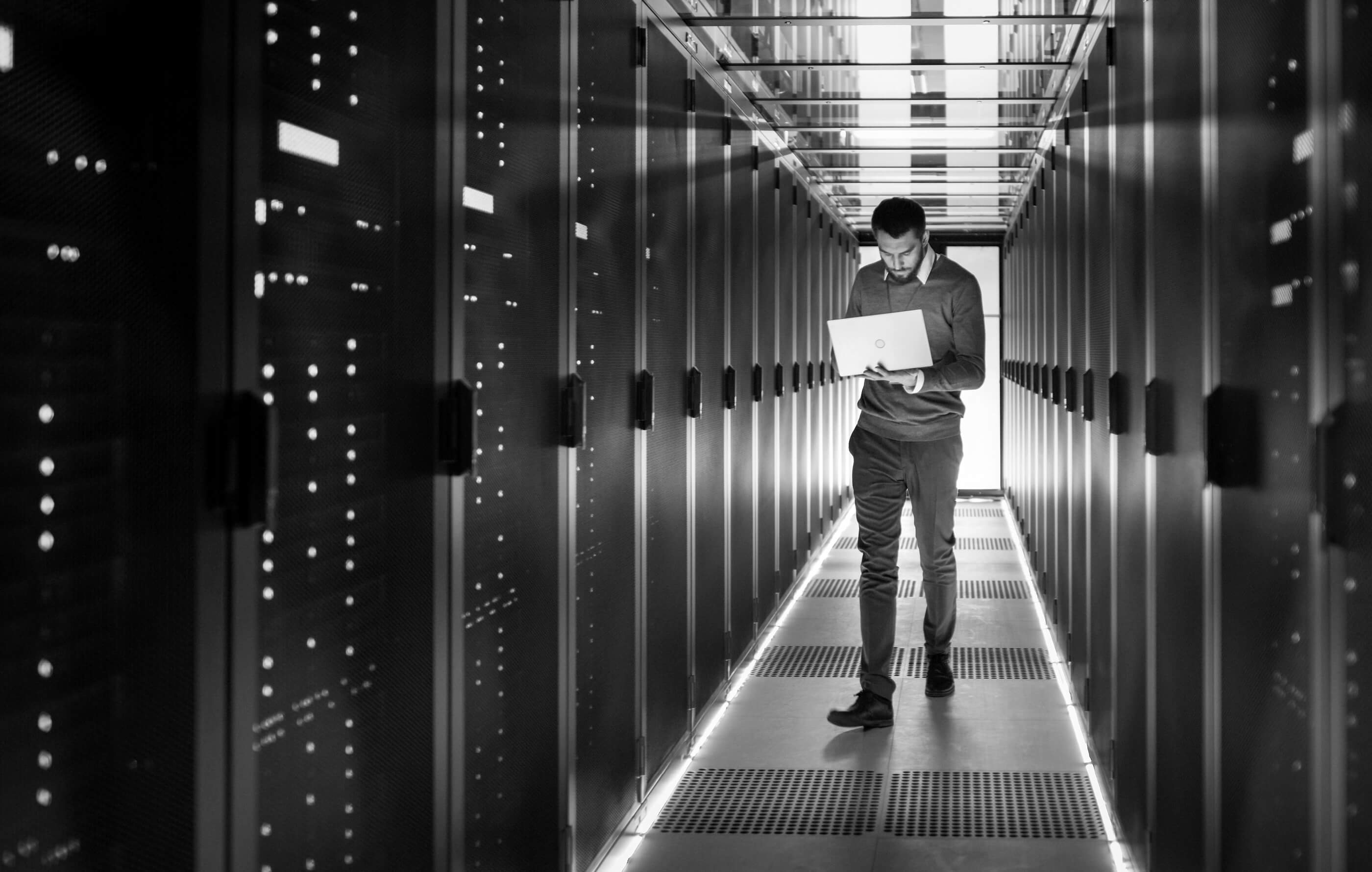 Full screen black and white image of a man walking along a server corridor, holding a laptop and looking at the screen