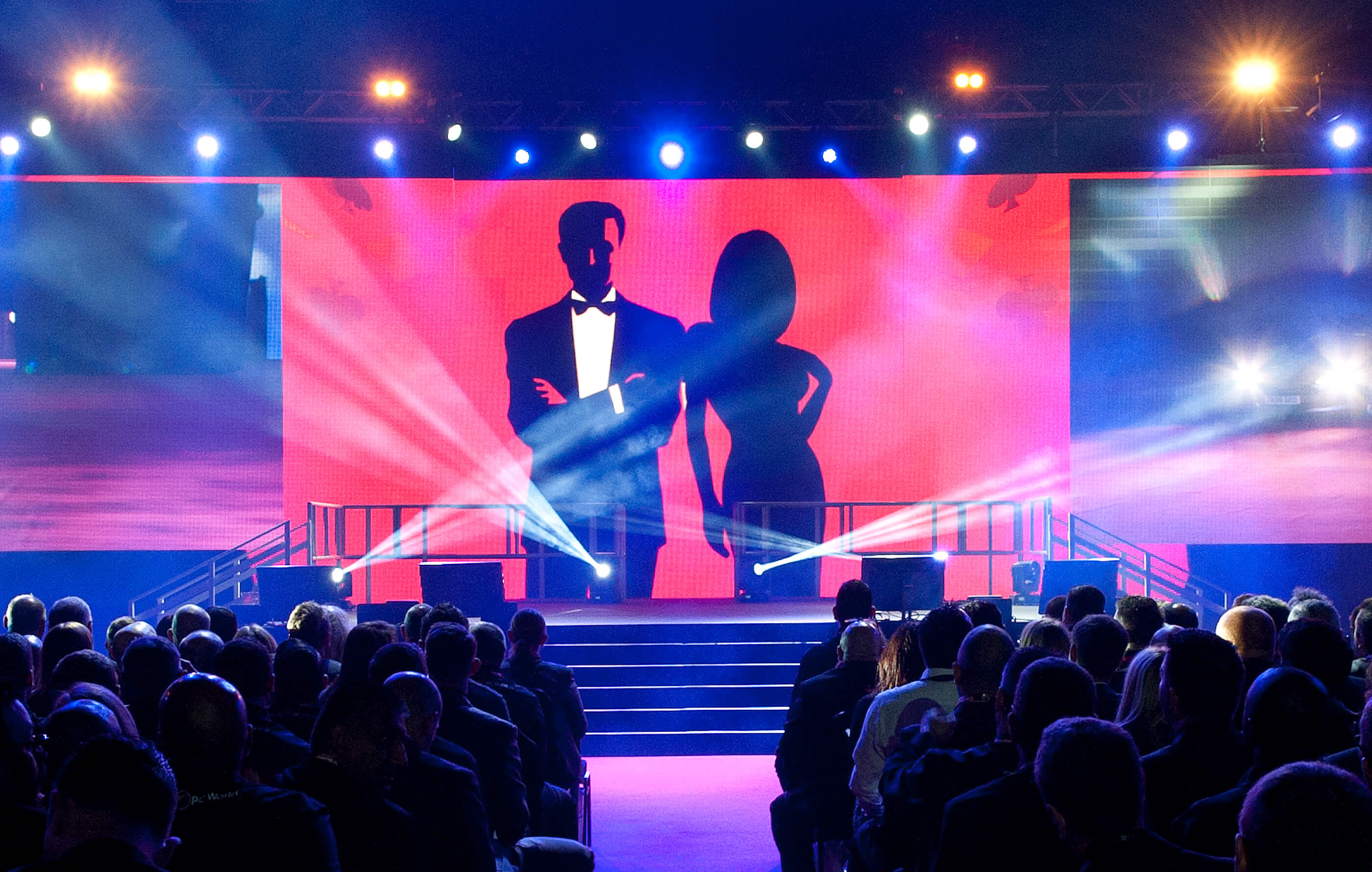 Photo from the event, taken from the back of the room facing the stage. The audience sit facing a red digital backdrop, with illustrations of a male and female silhouette in the centre
