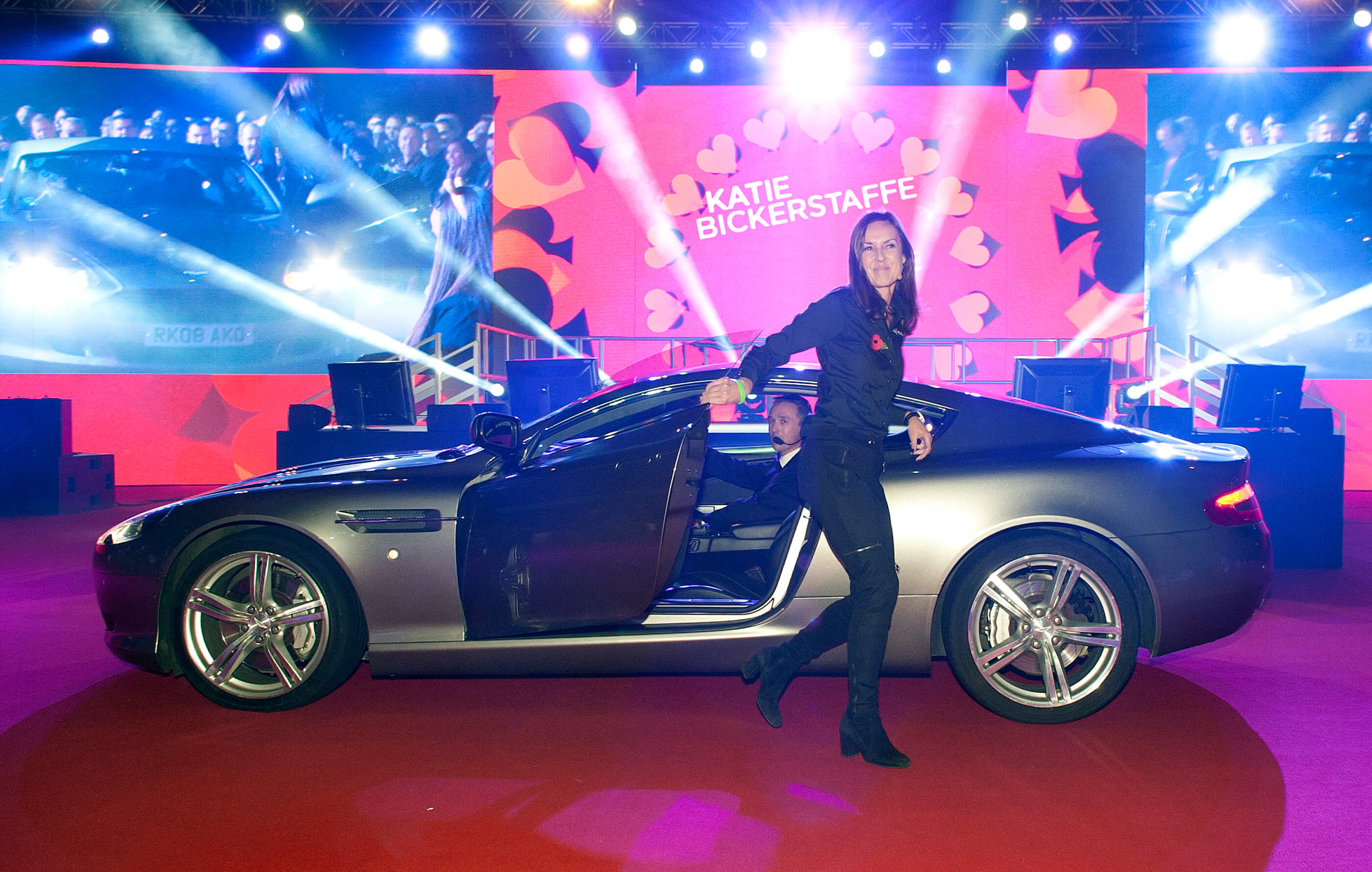 Katie Bickerstaffe – Chief Executive of Dixons UK and Ireland – steps out of a grey Aston Martin DB7 in front of the main stage