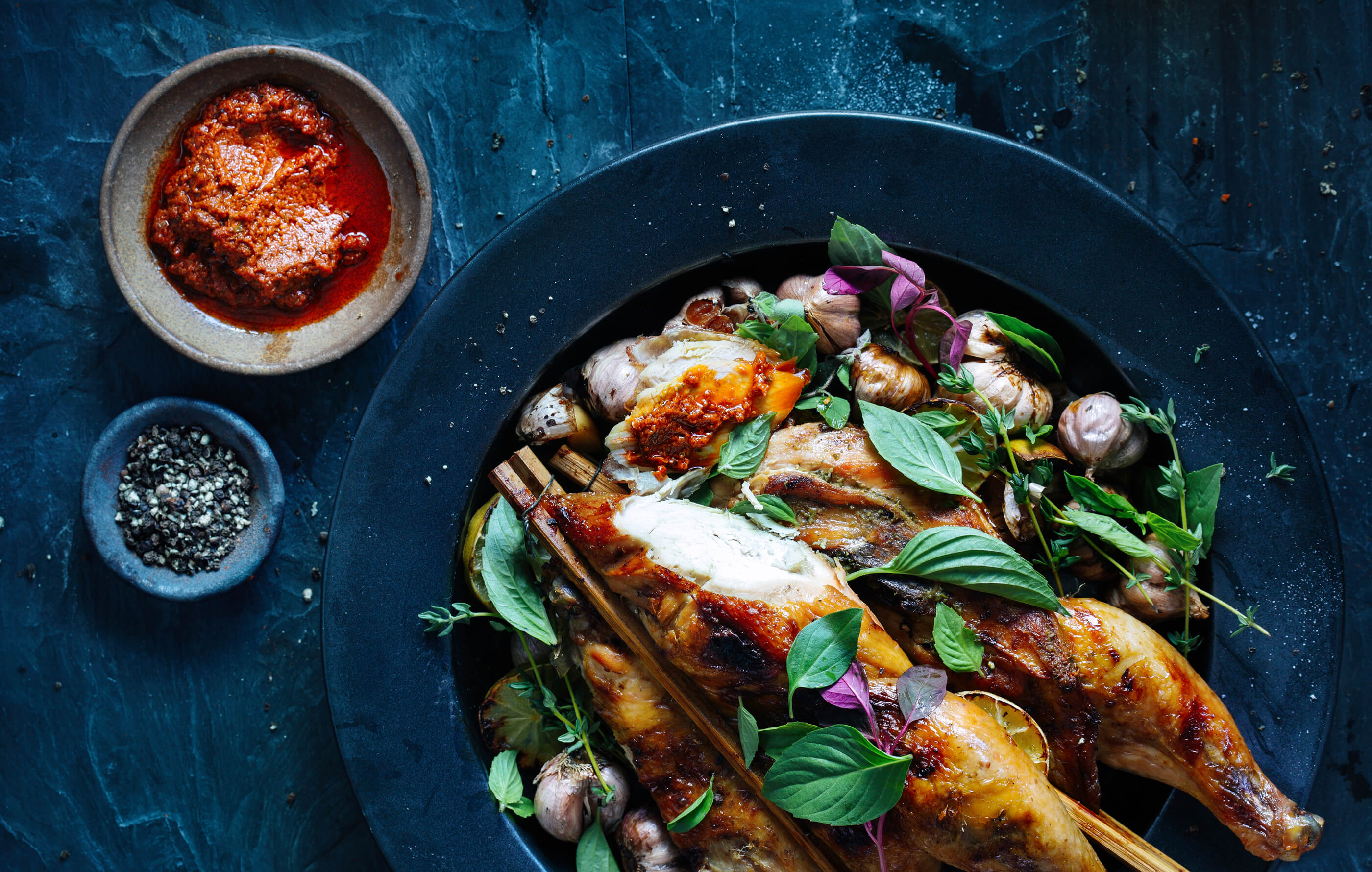 Full spread image of roasted chicken in a dark blue bowl, with roasted garlic, rosemary and a sprinkle of fresh herbs
